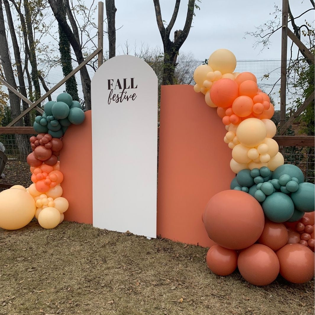 Outdoor fall-themed balloon garland backdrop with a white arch sign reading 'FALL festive' on a terracotta and peach wall, decorated with orange, peach, cream, and teal balloons on grass.